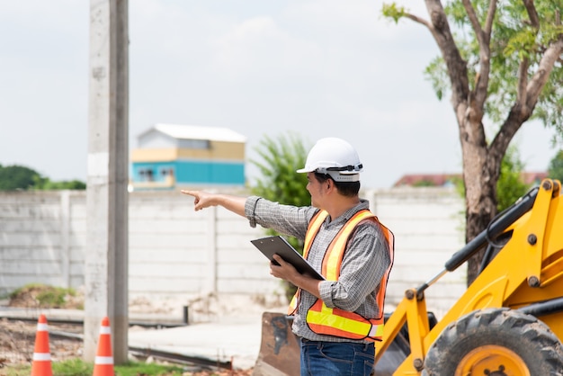 Asian man civil construction engineer worker or architect with helmet and safety vest working and holding a paper board note for see blueprints or plan at a building or construction site