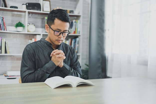 Asian man doing hands together in prayer to God along with the bible In the Christian concept of faith spirituality and religion men pray in the Bible prayer bible in the living room at home