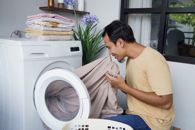 asian Man doing laundry at home loading clothes into washing machine smell the linen