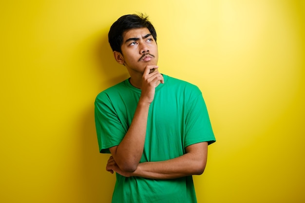 Asian man in green t-shirt thinking something, looking for good idea against yellow background