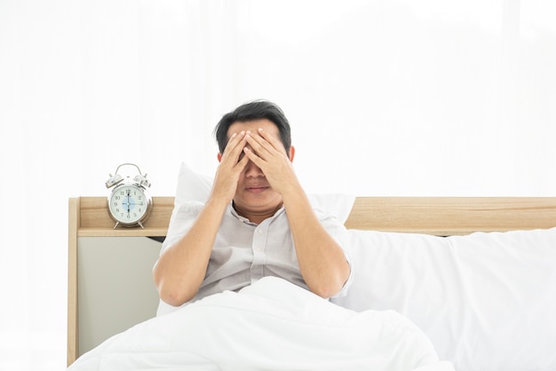 Asian man hand cover face while sit on bed with alarm clock at 6 am.