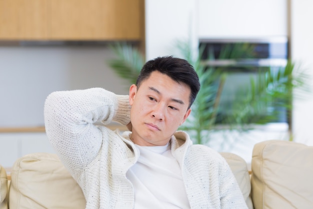 asian man holding his head with a severe headache at home in a room on the couch