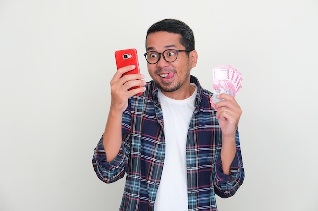 Asian man looking to his handphone with funny face expression while holding money