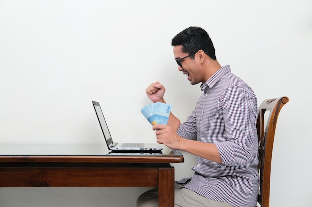 Asian man looking to his laptop while holding money with excited expression