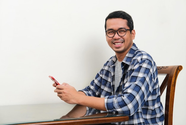 Asian man looking and smiling to the camera while using mobile phone