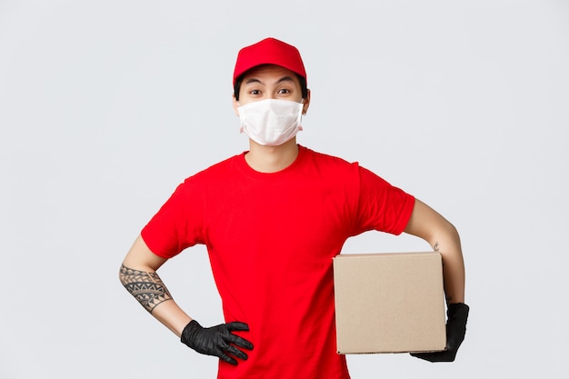 Asian man in red cap and t-shirt, holding parcel box, wear protective gloves and medical mask. Courier pick-up package from client, shipping box during covid-19 pandemic outbreak