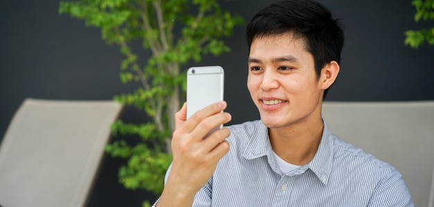 asian man resting at garden outdoor holding smartphone to watching multimedia on internet network and smiling with satisfying feeling