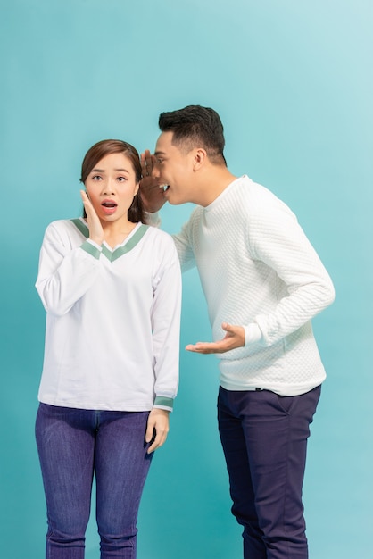 Asian man sharing secret or whispering gossips into his girlfriend's ear, blue studio