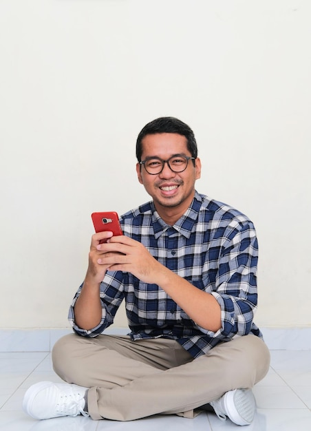 Asian man sitting in the floor smiling to camera while using his handphone