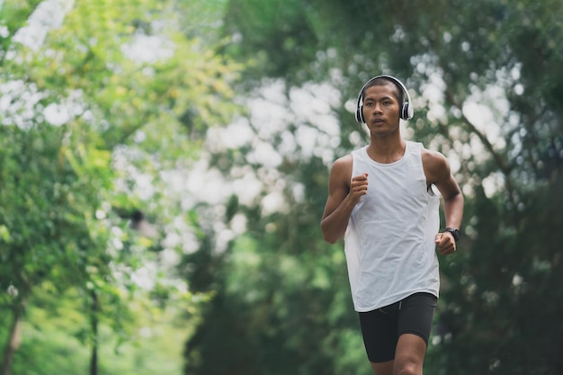 Asian man sportsman fit body wearing white headphone and smart watch while running or jogging in the garden have more tree and clean air exercising at park Sport healthy running concept