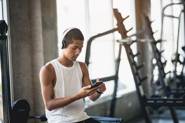 Asian man sportsman fit body wearing white headphone and using mobile phone to listen music while resting or before running exercising at fitness gym Sport healthy running fitness gym concept