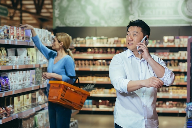 An asian man in a supermarket makes purchases chooses products and talks on the phone consults with