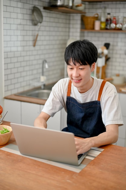 Asian man using laptop in his kitchen joining online cooking class searching food recipe