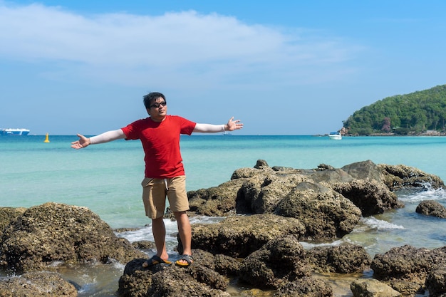 Asian man wear red Tshirt standing on rock open hands feel freedom and relax on blue sea beach in sunny day