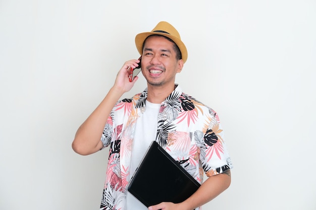 Asian man wearing beach shirt holding laptop and aswering a phone call with happy expression