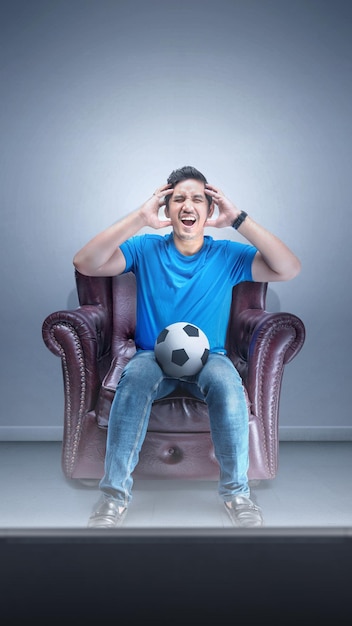 Asian man with the ball sitting on the couch with a disappointed expression watching football