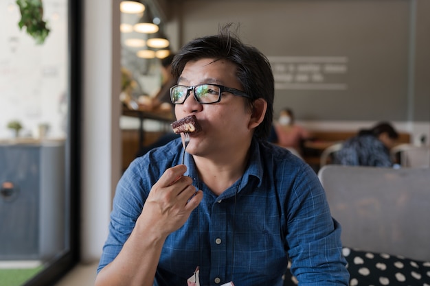 Asian man with eyeglasses eating chocolate brownie in cafe looking outside, man holding folk with brownie cake putting in mouth