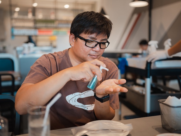 Asian man with eyeglasses using alcohol gel from small bottle to clean hands and protect corona virus or COVID-19 infection before eating in restaurant