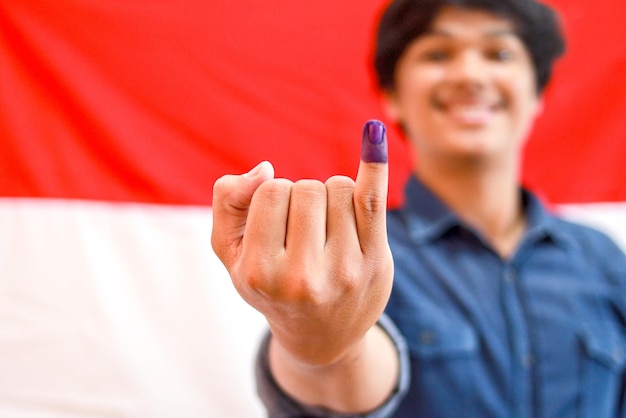 Asian man with Indonesia flag on the background showing little finger with blue ink patches as a mar