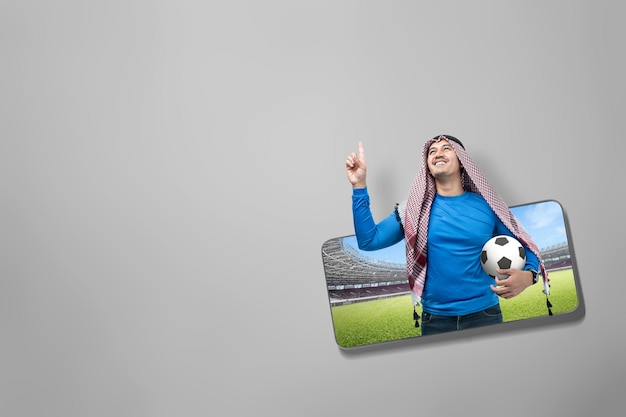 Asian man with keffiyeh standing while holding the ball with an excited expression on the football stadium