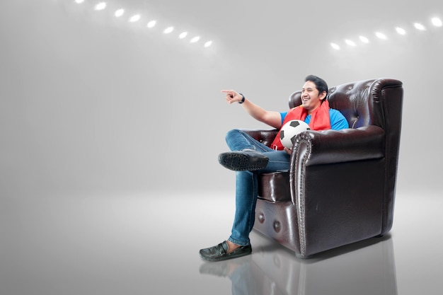 Asian man with scarf and ball sitting on the couch watching football