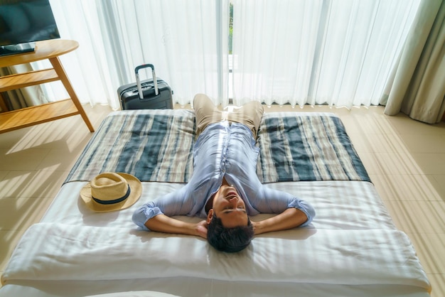 Asian man with a suitcase lay down on a bed in his room in resort after his arrival during his summer vacation holiday travel.