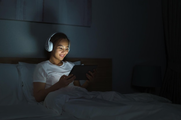 Asian mobile addict woman using digital tablet browsing the internet for watching movie or virtual meeting video call online with boyfriend on the bed before she sleeping at night.