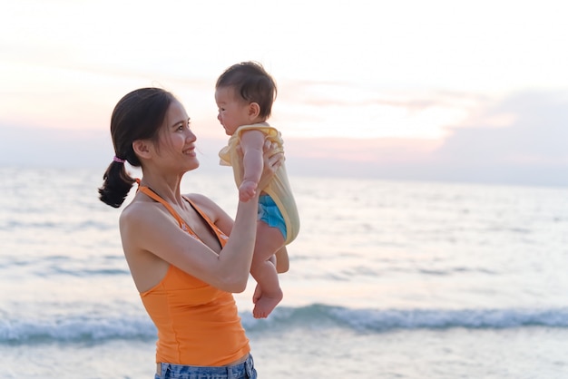 Asian mother standing on the beach holding her baby in two arms raising the kid and looking at her child.