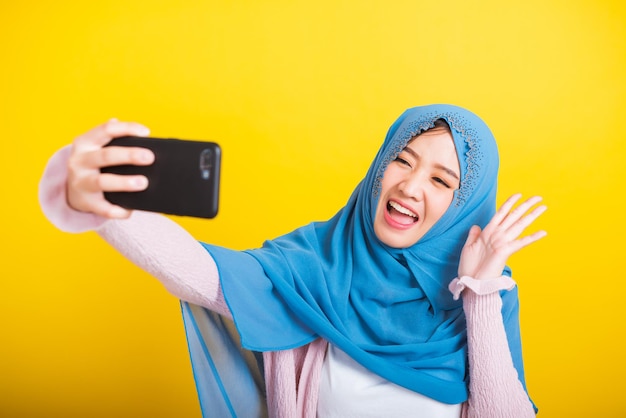 Asian Muslim Arab, Portrait of happy beautiful young woman Islam religious wear veil hijab funny smile she taking making selfie with smart mobile phone, studio shot isolated on yellow background