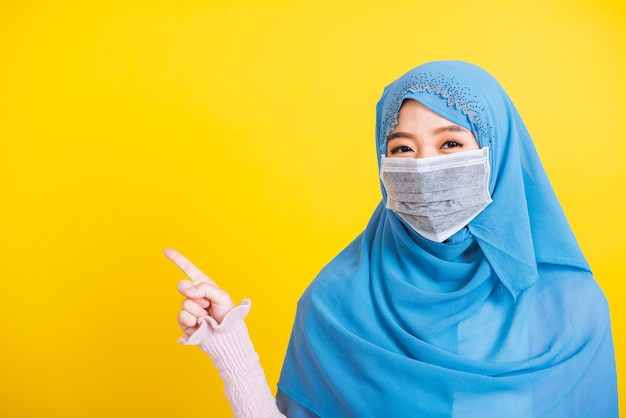 Asian Muslim Arab, Portrait of happy beautiful young woman religious wear veil hijab and face mask protective to prevent coronavirus she pointing finger side away space, isolated on yellow background