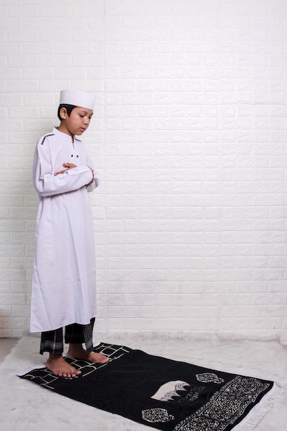 Asian muslim kid in white shirt, sarong and skullcap stands with hand in chest, praying salah on mat