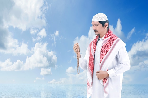 Asian Muslim man praying with prayer beads on his hands with a blue sky