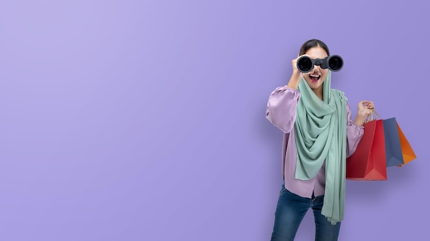 Asian Muslim woman in a headscarf looking through binoculars while holding shopping bags