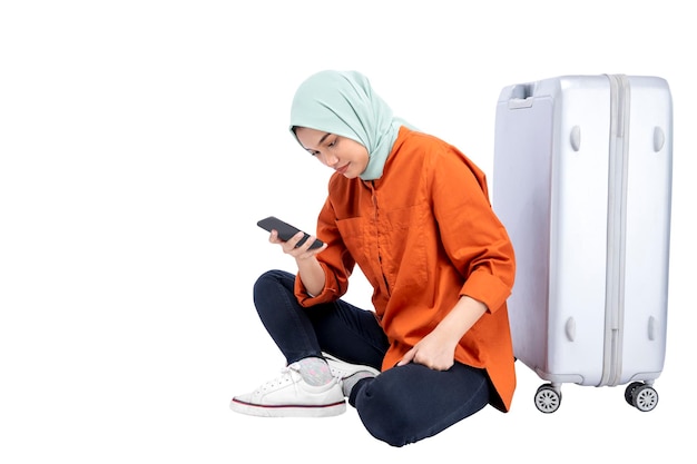 Asian Muslim woman in a headscarf sitting with a suitcase while using a mobile phone