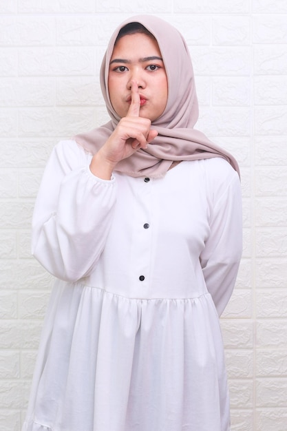 Asian Muslim woman in hijab showing hush sign putting finger on lips gesturing keep silence