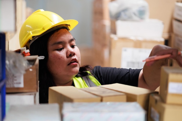 Asian obese female warehouse worker in safety vest and hardhat helmet in warehouse factory industrial product in cardboard boxes on shelves Plus size female inspecting products while working