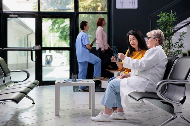 Asian patient analyzing radiography scan with physician specialist in medical clinic lobby, looking at bones x ray exam results to find disease diagnosis. Doing healthcare consultation appointment.