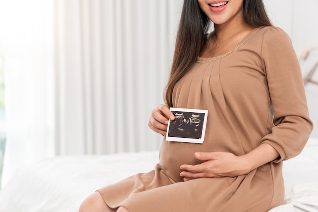 Asian pregnant woman in brown dress holding ultrasound on her belly in bed at bedroom. Pregnancy, parenthood, preparation and expectation concept