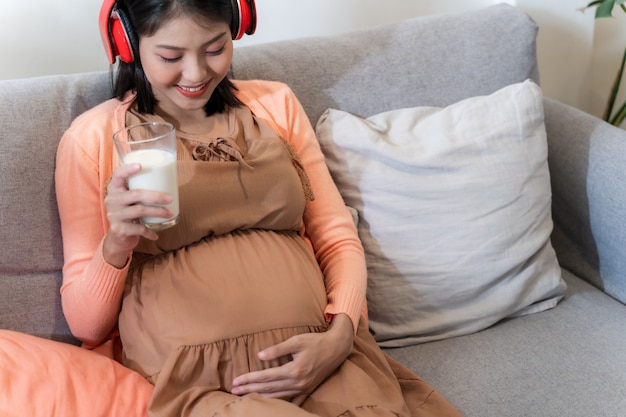 An Asian pregnant woman smile and sitting on the sofa and listening to music and hold a glass of milk with feeling happily and relaxed.