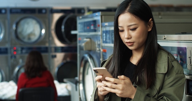 Asian pretty woman with long dark hair tapping and texting message on smartphone while standing in laundry service room. Beautiful woman typing on phone and waiting for clothes to wash.