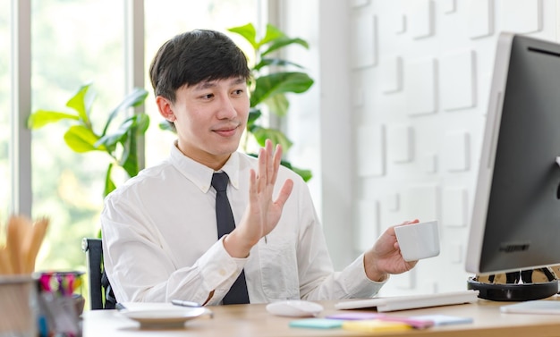 Asian professional successful male businessman employee in formal shirt with necktie sitting hold coffee cup waving hand greeting say hi with colleague in computer monitor at working desk in office.