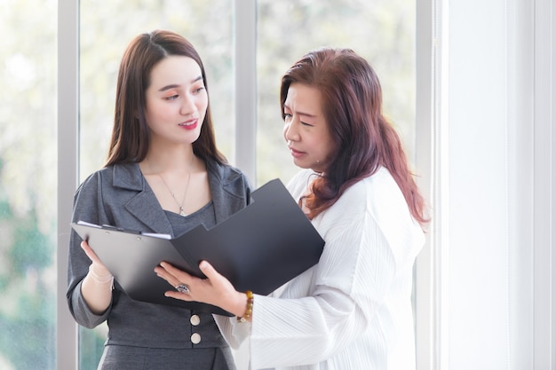 Asian professional woman talks with her woman boss to consult about work in file document in her hands at workplace.