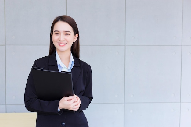 Asian professional working woman in black suit holds clipboard in her hands and confident smiles