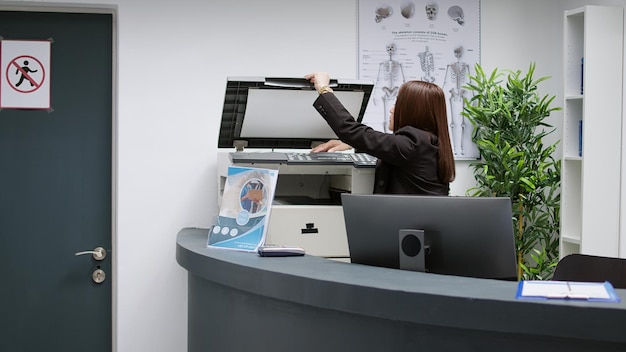 Asian receptionist working with medical reports at reception counter desk in hospital lobby waiting room. Employee helping with healthcare appointments and consultations, clinical center.