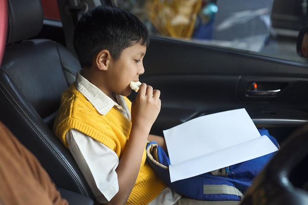 Asian school boy wearing uniform is sitting in a car seat reading a book while eating bread