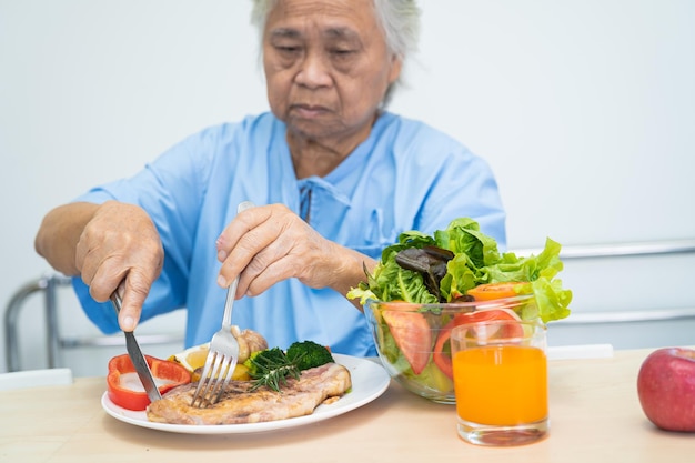 Asian senior or elderly old lady woman patient eating breakfast and vegetable healthy food with hope and happy while sitting and hungry on bed in hospital