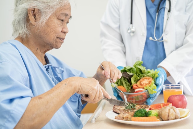 Asian senior or elderly old lady woman patient eating breakfast and vegetable healthy food with hope and happy while sitting and hungry on bed in hospital