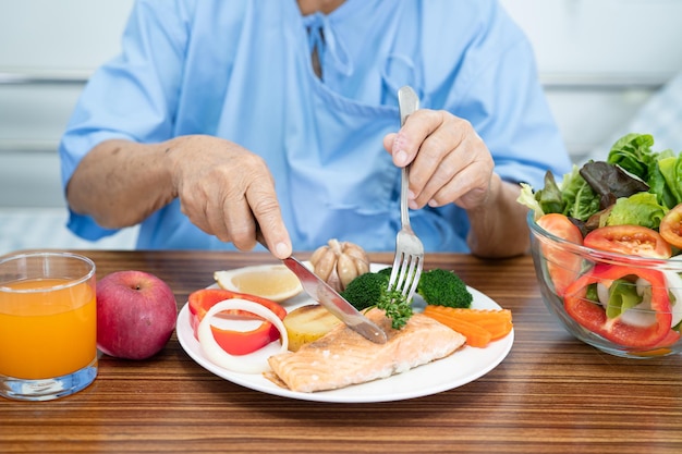 Asian senior or elderly old lady woman patient eating Salmon steak breakfast with vegetable healthy food while sitting and hungry on bed in hospital