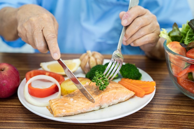 Asian senior or elderly old lady woman patient eating Salmon steak breakfast with vegetable healthy food while sitting and hungry on bed in hospital