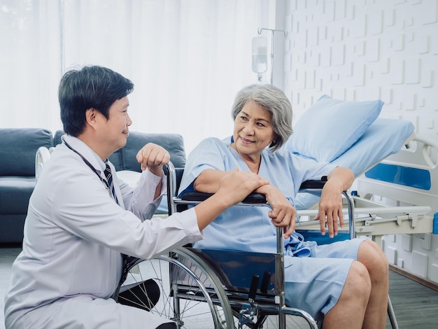 Asian senior woman patient dressed in blue smiling happily sits in wheelchair with kindly man doctor in white suit holding her hand beside in recovery room in hospital Healthcare support concept